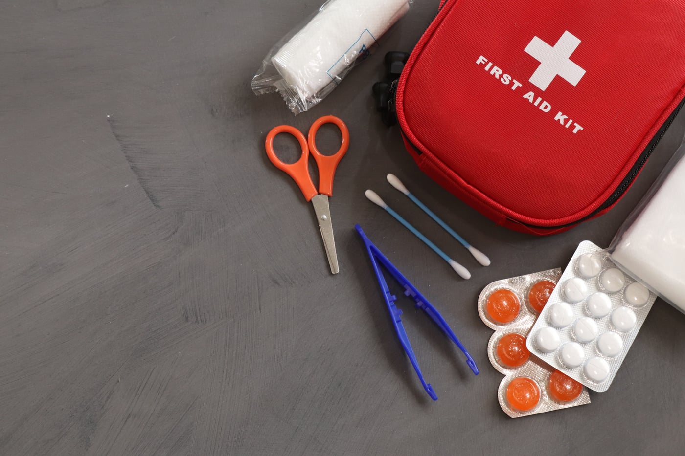 A red first aid kit with scissors, tweezers, cotton swabs and blister packs of tablets on a dark table