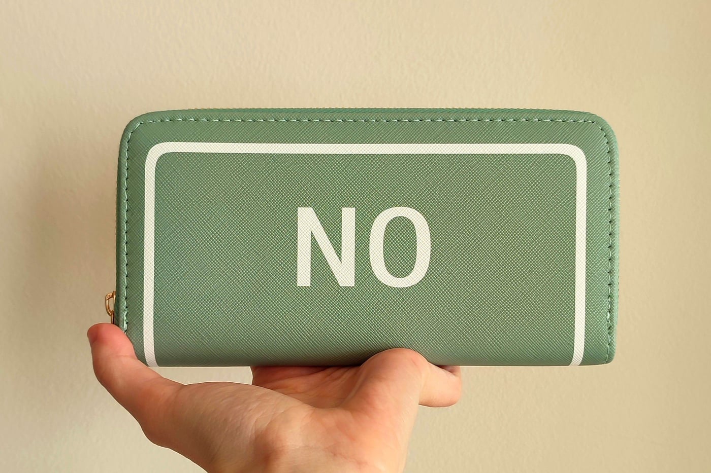 A hand holding up a green wallet printed with the word NO against a plain wall