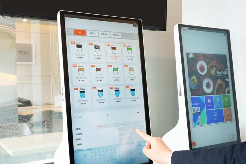 A Korean cafe self-service kiosk with an English-style menu grid of hot and iced coffees and a hand tapping the touch screen